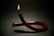 A unique candle with a deep red color is twisted into a loop, with its wick lit and burning steadily. The background is dark, enhancing the candle's bright flame and glossy surface. Wax drips can be seen on the surface where the candle rests.