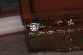 A sparkling diamond ring rests on the edge of a wooden jewelry box with ornate metal detailing. The focus is on the ring, highlighting its intricate design and shine. Soft lighting creates a warm, intimate atmosphere.