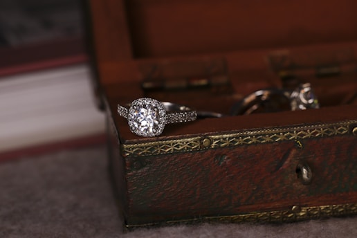 A sparkling diamond ring rests on the edge of a wooden jewelry box with ornate metal detailing. The focus is on the ring, highlighting its intricate design and shine. Soft lighting creates a warm, intimate atmosphere.