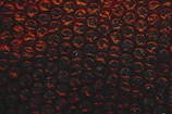A close-up view of a sheet of bubble wrap with a dark, dramatic lighting. Each bubble is illuminated with an intense red and black contrast, highlighting the texture and translucency of the material.