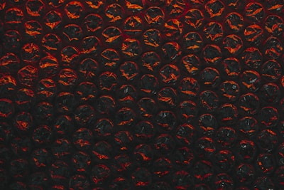 A close-up view of a sheet of bubble wrap with a dark, dramatic lighting. Each bubble is illuminated with an intense red and black contrast, highlighting the texture and translucency of the material.