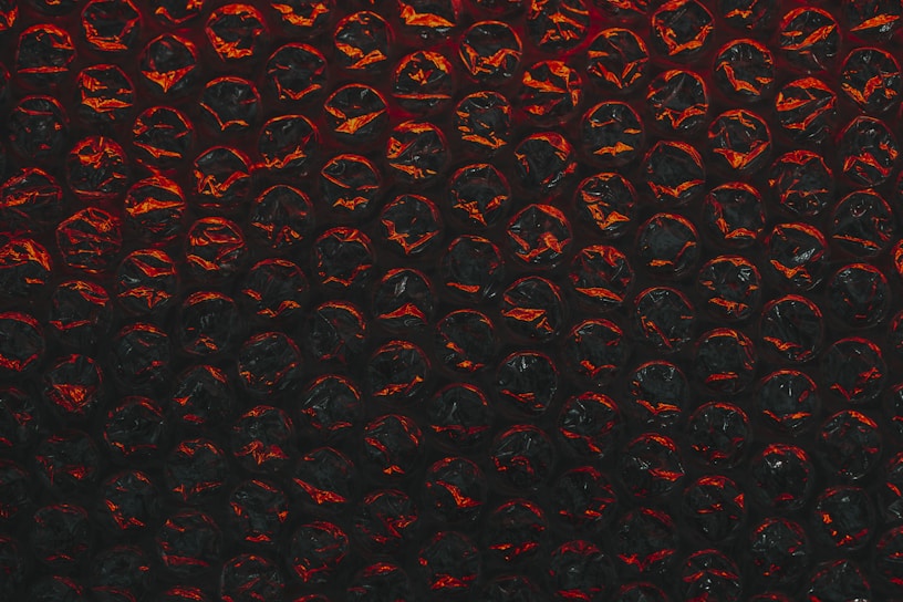 A close-up view of a sheet of bubble wrap with a dark, dramatic lighting. Each bubble is illuminated with an intense red and black contrast, highlighting the texture and translucency of the material.