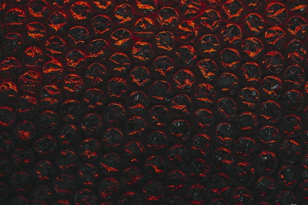 A close-up view of a sheet of bubble wrap with a dark, dramatic lighting. Each bubble is illuminated with an intense red and black contrast, highlighting the texture and translucency of the material.