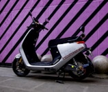 A modern electric scooter is parked against a vibrant purple wall with black diagonal stripes. The scooter has a sleek black and white design, with a comfortable seat and handlebars equipped with mirrors. The ground is paved, and there are decorative concrete spheres near the wall.