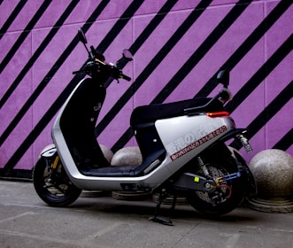 A modern electric scooter is parked against a vibrant purple wall with black diagonal stripes. The scooter has a sleek black and white design, with a comfortable seat and handlebars equipped with mirrors. The ground is paved, and there are decorative concrete spheres near the wall.