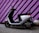 A modern electric scooter is parked against a vibrant purple wall with black diagonal stripes. The scooter has a sleek black and white design, with a comfortable seat and handlebars equipped with mirrors. The ground is paved, and there are decorative concrete spheres near the wall.