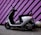 A modern electric scooter is parked against a vibrant purple wall with black diagonal stripes. The scooter has a sleek black and white design, with a comfortable seat and handlebars equipped with mirrors. The ground is paved, and there are decorative concrete spheres near the wall.
