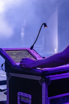 A person using a touch screen device that is mounted on a large, sturdy console or flight case. There is a flexible lamp attached to the device, providing some illumination on the screen. The background is filled with a smoky or foggy atmosphere, typically seen at events or concerts.