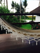 A serene outdoor scene featuring a neatly placed hammock on a wooden deck overlooking a lush green garden. The garden is adorned with palm trees and vibrant red flowers, and there's a glimpse of a white building with a thatched roof. The sky peeks through the trees in the background.