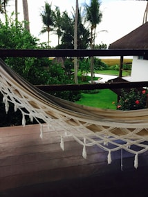 A serene outdoor scene featuring a neatly placed hammock on a wooden deck overlooking a lush green garden. The garden is adorned with palm trees and vibrant red flowers, and there's a glimpse of a white building with a thatched roof. The sky peeks through the trees in the background.