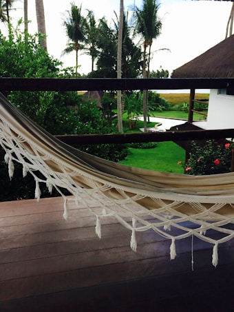 A serene outdoor scene featuring a neatly placed hammock on a wooden deck overlooking a lush green garden. The garden is adorned with palm trees and vibrant red flowers, and there's a glimpse of a white building with a thatched roof. The sky peeks through the trees in the background.