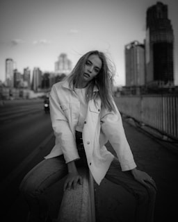 A black-and-white photograph featuring a person sitting on a railing in an urban setting. The individual has long hair and is wearing a light-colored jacket and jeans. The city skyline with tall buildings is blurred in the background, adding depth to the scene.