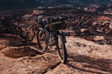 A heavily equipped mountain bike stands on a rocky cliff, overlooking an expansive canyon with layers of red and brown rock formations. Bushes sparsely dot the rugged landscape, and the scene is bathed in warm, late afternoon light.