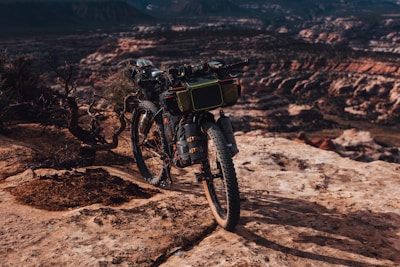 A heavily equipped mountain bike stands on a rocky cliff, overlooking an expansive canyon with layers of red and brown rock formations. Bushes sparsely dot the rugged landscape, and the scene is bathed in warm, late afternoon light.