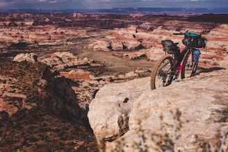 A loaded mountain bike is parked on the edge of a rocky cliff, overlooking a vast desert landscape with layered rock formations and a mix of shadows and sunlight.