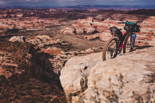 A loaded mountain bike is parked on the edge of a rocky cliff, overlooking a vast desert landscape with layered rock formations and a mix of shadows and sunlight.