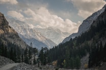 A scenic mountain landscape with a rugged pathway winding through a valley. Snow-capped peaks rise majestically in the background, surrounded by dense forests of pine trees. The sky is partially covered with fluffy clouds, creating a serene and picturesque atmosphere.