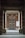 An entrance framed by two large stone pillars leading up to a wooden door with a geometric pattern. The surrounding walls are made of red brick, and there is a blank white board mounted on the right side. A small stone step leads to the entrance area, creating a sense of symmetry.