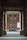 An entrance framed by two large stone pillars leading up to a wooden door with a geometric pattern. The surrounding walls are made of red brick, and there is a blank white board mounted on the right side. A small stone step leads to the entrance area, creating a sense of symmetry.