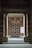 An entrance framed by two large stone pillars leading up to a wooden door with a geometric pattern. The surrounding walls are made of red brick, and there is a blank white board mounted on the right side. A small stone step leads to the entrance area, creating a sense of symmetry.