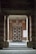 An entrance framed by two large stone pillars leading up to a wooden door with a geometric pattern. The surrounding walls are made of red brick, and there is a blank white board mounted on the right side. A small stone step leads to the entrance area, creating a sense of symmetry.