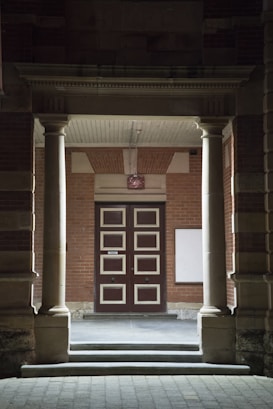 An entrance framed by two large stone pillars leading up to a wooden door with a geometric pattern. The surrounding walls are made of red brick, and there is a blank white board mounted on the right side. A small stone step leads to the entrance area, creating a sense of symmetry.