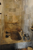 A stone bathroom features a built-in bathtub with antique stone and ceramic vessels placed around its edges. The walls are lined with tiles, and a fabric hangs over a rod on the far wall, adding a rustic and historical atmosphere.