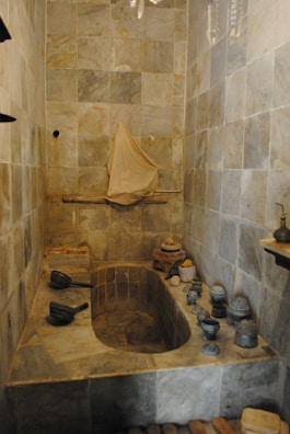 A stone bathroom features a built-in bathtub with antique stone and ceramic vessels placed around its edges. The walls are lined with tiles, and a fabric hangs over a rod on the far wall, adding a rustic and historical atmosphere.