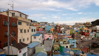 A vibrant hillside community with a multitude of colorful buildings stacked closely together. The structures range in color, featuring blues, pinks, reds, greens, and yellows, creating a visually striking contrast against the cloudy sky. Electrical lines and poles are visible, adding a layer of complexity to the urban landscape.
