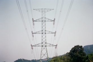 A tall metal electricity pylon stands prominently against a clear sky, with multiple power lines extending outward. Trees and hills are visible in the background, suggesting a rural or semi-rural setting.