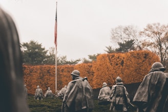 A group of military statues depicting soldiers wearing cloaks and helmets march in formation. An American flag is visible in the background, standing tall amidst autumn trees with orange and brown foliage.