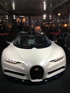 A sleek, white luxury sports car with an aerodynamic design is displayed prominently inside a glass-walled showroom. The car's headlights are styled with LED lights, and the vehicle features the emblem of a high-end car manufacturer on the front grille. Reflections of city lights and a bustling street scene are visible in the background.