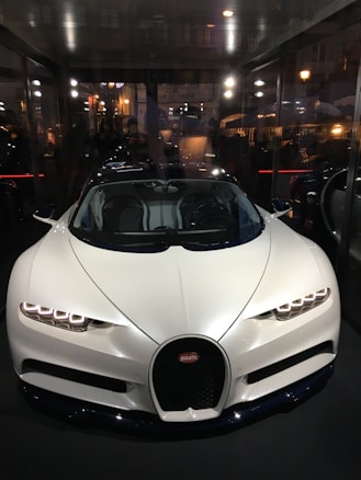 A sleek, white luxury sports car with an aerodynamic design is displayed prominently inside a glass-walled showroom. The car's headlights are styled with LED lights, and the vehicle features the emblem of a high-end car manufacturer on the front grille. Reflections of city lights and a bustling street scene are visible in the background.