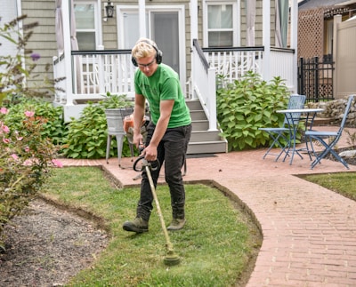 A person wearing protective headphones and glasses is using a grass trimmer in a well-maintained garden. The surroundings include a patch of green grass, a brick pathway, and a background with a house featuring a small porch, white railing, and large windows. There are hydrangea bushes and a blue outdoor chair and table set near the walkway.
