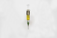 A syringe filled with a golden liquid, labeled 'The Clear Golden Goat', is centered on a white background. The syringe features measurement markings and a green tip.