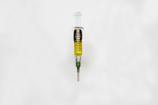 A syringe filled with a golden liquid, labeled 'The Clear Golden Goat', is centered on a white background. The syringe features measurement markings and a green tip.