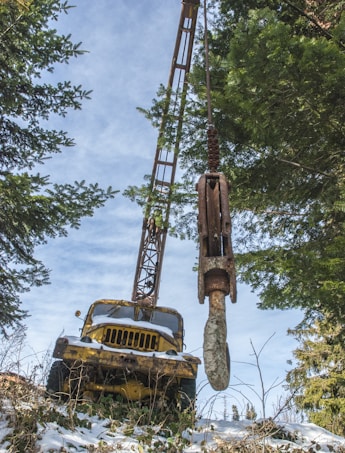 A yellow crane truck is positioned on a snowy, inclined forest path. The crane is extended upwards, with a large metal hook suspended in front. Surrounding the vehicle are tall evergreen trees, and patches of snow cover the ground and parts of the truck.