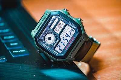 A digital wristwatch with a green casing is placed on a dark surface near a partially visible keyboard. The watch displays the time, features various function labels such as world time, illuminator, and has a detailed digital interface with multiple sections.