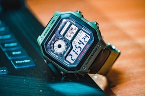 A digital wristwatch with a green casing is placed on a dark surface near a partially visible keyboard. The watch displays the time, features various function labels such as world time, illuminator, and has a detailed digital interface with multiple sections.