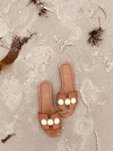 Two brown sandals with decorative white circles rest on a sandy beach. Seaweed and small pieces of driftwood are scattered nearby on the textured sand.