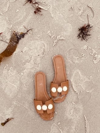 Two brown sandals with decorative white circles rest on a sandy beach. Seaweed and small pieces of driftwood are scattered nearby on the textured sand.