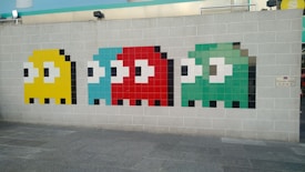 Mosaic art on a grey brick wall depicting four stylized ghost characters in pixelated form. Each ghost is distinct in color: yellow, light blue, red, and green, and they are aligned horizontally. The artwork reflects a retro video game style, reminiscent of classic arcade games. A small sign on the wall reads 'Private Area' in both English and Chinese script.