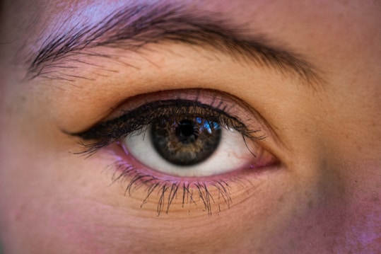 A close-up photograph of a human eye with detailed features, including well-defined eyelashes, an eyeliner, and a visible eyebrow. The iris is showcased with intricate patterns and color variations.