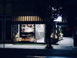 A small outdoor fruit and vegetable shop is softly illuminated at night. The storefront displays a variety of produce, including apples, oranges, and other fresh fruits, organized neatly in baskets. The ambient light highlights the entrance and the shop sign, while the surrounding area remains shrouded in darkness, except for some faintly visible greenery.