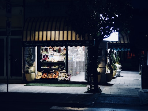 A small outdoor fruit and vegetable shop is softly illuminated at night. The storefront displays a variety of produce, including apples, oranges, and other fresh fruits, organized neatly in baskets. The ambient light highlights the entrance and the shop sign, while the surrounding area remains shrouded in darkness, except for some faintly visible greenery.