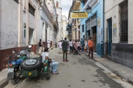 A narrow street scene with a group of people gathered near La Bodeguita Del Medio. The vibrant buildings are adorned with graffiti and posters. A green motorcycle with a sidecar is parked on the side, and people are casually dressed, seemingly engaged in conversation or strolling.