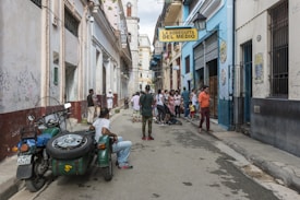 A narrow street scene with a group of people gathered near La Bodeguita Del Medio. The vibrant buildings are adorned with graffiti and posters. A green motorcycle with a sidecar is parked on the side, and people are casually dressed, seemingly engaged in conversation or strolling.