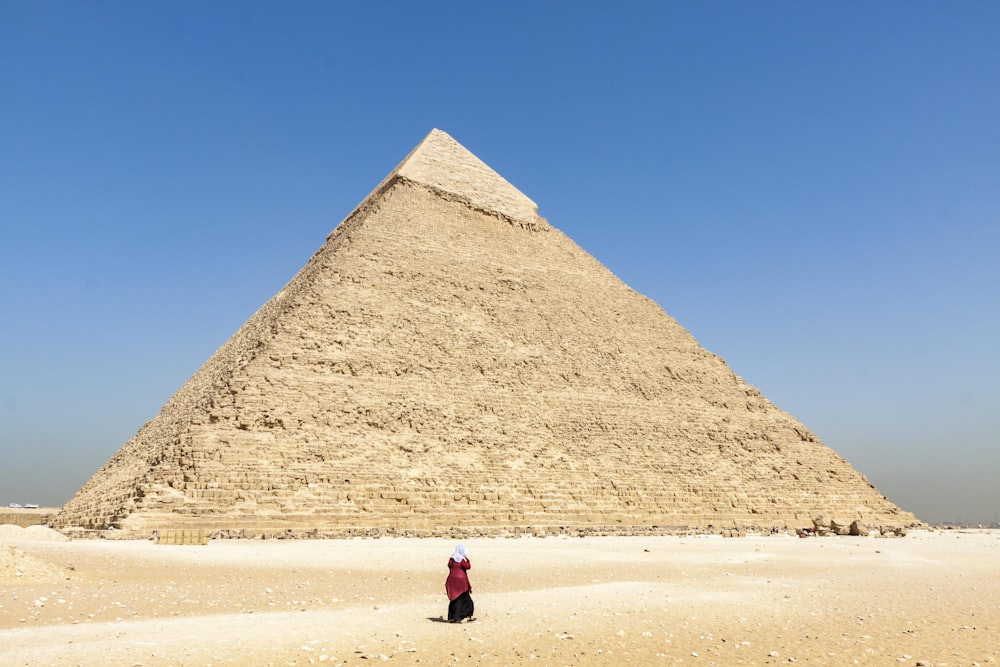 woman in black jacket walking on brown sand near pyramid under blue sky during daytime