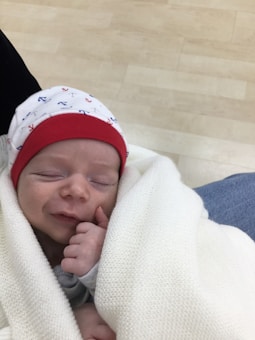 A newborn is peacefully sleeping while being swaddled in a soft, white blanket. A cozy hat with a red band and small anchor patterns covers the baby's head. The baby has a light smile and is gently holding a hand close to its mouth.