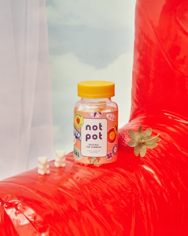 A bottle of CBD gummies labeled 'Not Pot' is placed on a red inflatable surface. The bottle features colorful, playful graphics. Two white bear-shaped gummies and a small green plant are nearby. The soft background includes a subtle view of clouds.